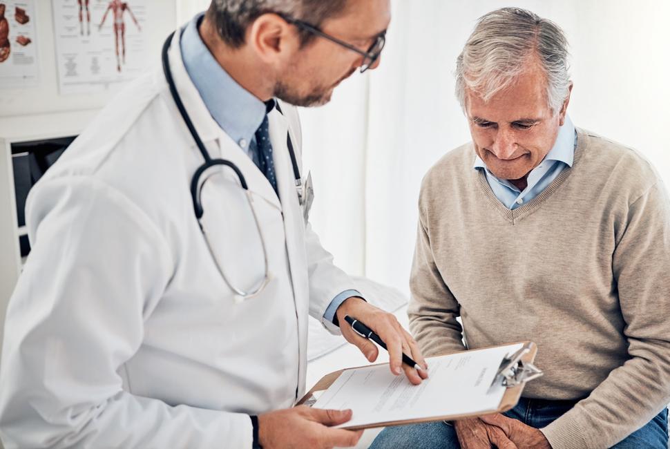 An older man in a v-neck sweater and button down shirt, seated, speaking with a doctor with glasses and a beard.