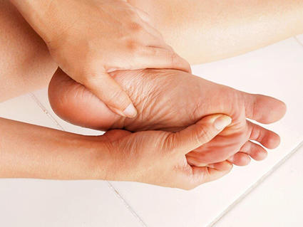 A close-up of a woman’s hands holding her foot. Her left hand ism mostly around the area of her heel; the right hand rubs the bottom of the foot below her big toe. A tile floor can be seen in the background.