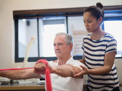 Physical therapist assisting an older male patient with resistance band exercises in a bright therapy clinic, with the patient stretching the red resistance band while the therapist provides guidance and support.